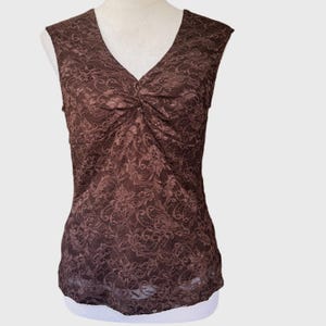 May include: A sleeveless brown top featuring an all-over floral lace pattern. The garment has a V-neckline with subtle gathered detailing at the center of the chest, creating a fitted silhouette that extends down to the straight hemline.