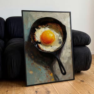 Puede incluir: Obra de arte enmarcada que representa un huevo frito en una sartén de hierro fundido negro. La yema es de color amarillo brillante, rodeada de clara, con un fondo oscuro. La pintura tiene un estilo de óleo texturizado.