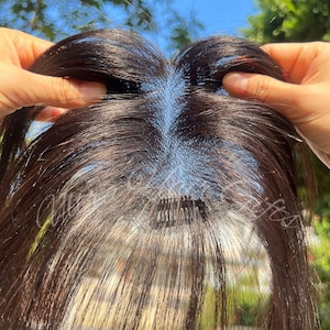 Op de afbeelding: Een donkerbruine haartop met een kanten basis, vastgehouden door handen. Het haarstuk heeft een natuurlijk ogende scheiding en is ontworpen om volume en dekking toe te voegen. Het haar is lang en steil.