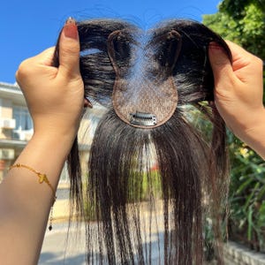 Puede incluir: Un postizo de cabello castaño oscuro con una pequeña base en forma de red y clips adjuntos. El cabello es largo y liso, con un aspecto natural. El postizo se sostiene contra un cielo brillante y despejado.