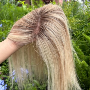 Può includere: Un topper per capelli biondi con radice castano scuro. I capelli sono lisci e hanno dei riflessi. Il topper è tenuto contro uno sfondo di fogliame verde e fiori blu.