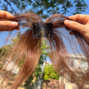 May include: A brown hairpiece with two black clips, held against a blurred background of greenery and sky. The hairpiece appears to be made of natural-looking hair fibers, designed to add volume or length to existing hair.