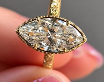 Op de afbeelding: Een gouden ring met een grote marquise-geslepen middensteen in een zetting met milgrain-rand. De band is gemaakt van geelgoud en is voorzien van een gedetailleerd, gegraveerd bloemmotief dat over de gehele oppervlakte doorloopt.