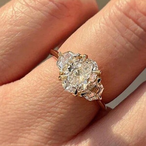 May include: A gold ring featuring a large, oval-cut diamond center stone flanked by two smaller, triangular-cut diamonds. The ring's band is a delicate gold color, and the diamonds sparkle brightly.