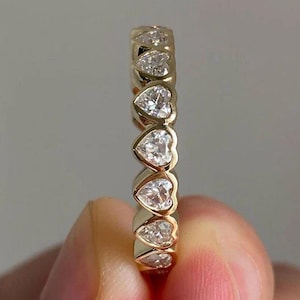 May include: A gold band ring featuring a series of heart-shaped, clear gemstones. The ring is held between fingers, showcasing the polished gold and the sparkle of the gemstones. The ring is a piece of jewelry.