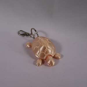 May include: A peach-colored turtle keychain with a silver-tone clasp. The turtle has a textured shell and four small flipper-like appendages. The keychain is a decorative accessory.