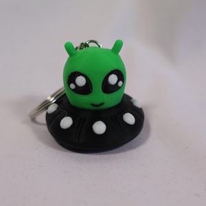 May include: A green alien keychain with large black eyes and white highlights. The alien has two small antennae and is attached to a black UFO-shaped base with white dots. The keychain is made of a smooth material and has a silver keyring.