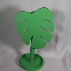 May include: A green, stylized monstera leaf decoration. The leaf is mounted on a stem and a circular base. The design is a solid, bright green color, and the background is white.