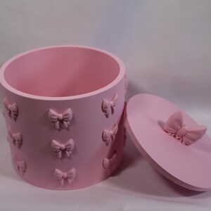 May include: A pale pink cylindrical container with a matching lid. The container features embossed bow details. The lid is round with a bow-shaped handle. The container is open, revealing a smooth interior.
