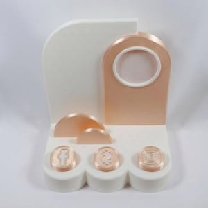 May include: A modern display stand in white and rose gold. The stand features a white base with three circular platforms holding rose gold objects. A rose gold arch with a circular opening and a white accent is centered.
