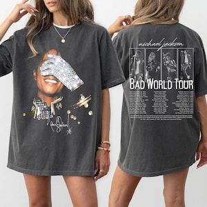 May include: Dark grey t-shirt with a graphic of Michael Jackson, a sparkling glove over his eye, and gold accents. The front has his signature. The back of the shirt features "Michael Jackson BAD WORLD TOUR" with tour dates and images.