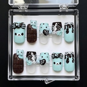 May include: A set of ten press-on nails in a clear plastic case. The nails feature various designs, including a bear with chocolate, musical notes, bows, and dripping chocolate. The base colours are light blue and clear, with brown and white accents.