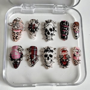 May include: A set of ten artificial nails with a punk rock aesthetic. The nails showcase diverse designs, such as skulls, leopard print, and tartan patterns, embellished with silver spikes and decorative elements. The nails are presented in a clear plastic case.