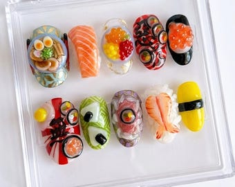 Könnte beinhalten: Eine Sammlung von Sushi-Nagelkunst-Designs in einer transparenten Vitrine. Die Nägel zeigen detaillierte Miniatur-Sushi, darunter Lachs, Rollen und Garnelen, mit leuchtenden Farben und Texturen. Die Designs sind aufwendig und kreativ.