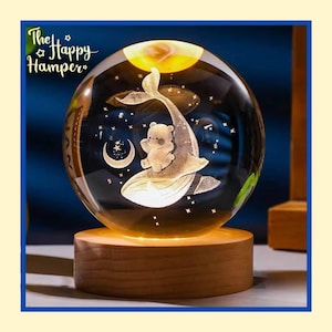 Puede incluir: Una esfera decorativa de cristal con un diseño de ballena y oso, una luna creciente y estrellas. La esfera está sobre una base de madera e iluminada desde dentro. El logotipo de The Happy Hamper es visible en la esquina superior izquierda.