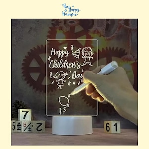 May include: An illuminated acrylic night light with the words "Happy Children's Day" and child illustrations. The light is on a white base and is being written on with a white marker. Wooden blocks with numbers are in the foreground. The Happy Hamper logo is at the top.