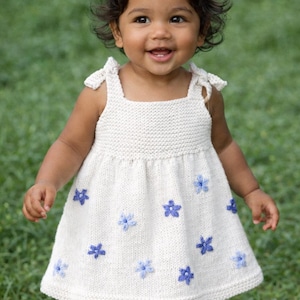 Puede incluir: Un vestido de punto color crema con detalles florales azules. El vestido tiene un escote cuadrado y tirantes anudados. Un lazo blanco adorna el pelo del niño. Adecuado para un bebé o un niño pequeño.
