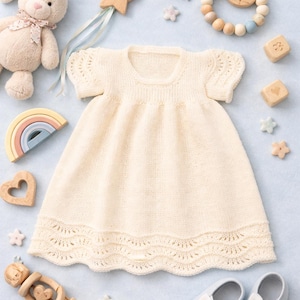 Könnte beinhalten: Cremefarbenes Babykleid mit kurzen Ärmeln und einem gewellten Saum, umgeben von Babyspielzeug. Ein Plüschhase, Holzspielzeug und Babyschuhe sind um das Kleid herum angeordnet. Das Kleid ist aus weichem Strickmaterial.