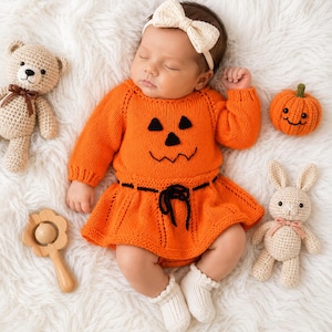 Puede incluir: Un bebé viste un vestido de punto naranja brillante de manga larga con un diseño de cara de calabaza negra y un lazo en la cintura. El bebé lleva una diadema con lazo color crema y calcetines blancos. Cerca hay un oso de peluche beige, una pequeña calabaza naranja, un sonajero de madera y un conejo de ganchillo color crema.
