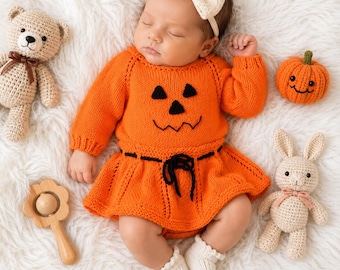 May include: A baby wearing a bright orange knitted long-sleeve dress with a black jack-o'-lantern face design and a black waist tie. The baby has a cream-colored bow headband and white socks. Nearby are a tan teddy bear, a small orange pumpkin, a wooden rattle, and a cream-colored crochet bunny.