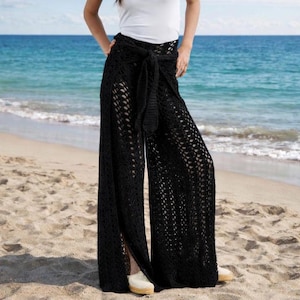 Op de afbeelding: Zwarte gehaakte broek met wijde pijpen en een strik in de taille. De broek heeft een split aan de voorkant en wordt gedragen op een zandstrand met de oceaan op de achtergrond. Het open ontwerp zorgt voor luchtcirculatie.