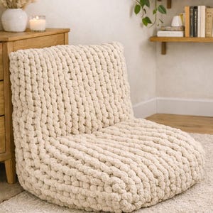 May include: A cream-colored, chunky knit floor chair with a rounded seat and a supportive backrest. The chair is made of thick, soft yarn and sits on a matching cream-colored rug. The chair is designed for comfortable lounging.