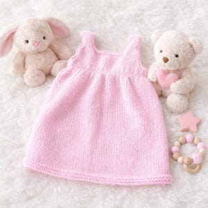 Può includere: Un vestito da neonato rosa chiaro lavorato a maglia con scollo tondo e gonna svasata. L'abito è circondato da un coniglietto di peluche, un orsacchiotto con un cuore rosa, un massaggiagengive a forma di stella e un massaggiagengive con perline in legno e rosa.