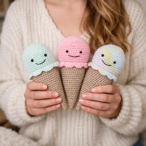 May include: Three handmade crochet ice cream cones in pastel colors. Each cone features a smiling face and a scalloped edge. The ice cream scoops are mint green, pink, and pastel yellow, atop brown cones.