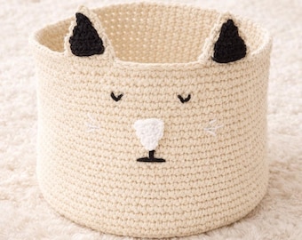 May include: A cream-colored, crocheted storage basket shaped like a cat. The basket features black ears, embroidered eyes, a white nose, and a black mouth. The round basket is designed for storing toys or other items.