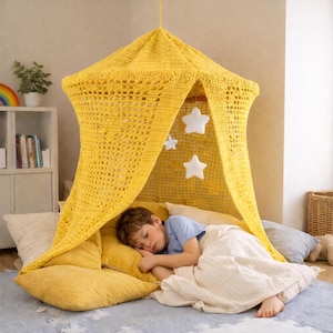May include: A yellow crochet canopy bed with star-shaped decorations hangs above a child sleeping on a bed of pillows. The canopy is suspended from the ceiling and provides a cozy, enclosed space. The bed is covered with a white blanket.