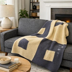 Puede incluir: Una manta con estampado geométrico azul marino y beige sobre un sofá gris. La manta presenta formas cuadradas y rectangulares grandes. El sofá está en un salón con una mesa de centro redonda de madera y una chimenea.