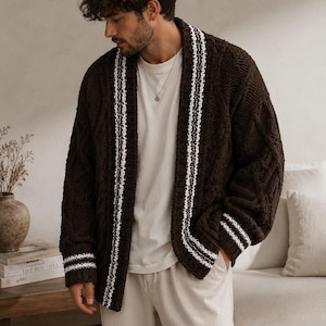 Peut inclure: Un cardigan en tricot torsadé marron foncé avec une bordure rayée blanche le long de l'ouverture avant et des poignets. Le vêtement est porté sur un t-shirt blanc uni et un pantalon clair, présentant un motif en tricot texturé sur les manches.