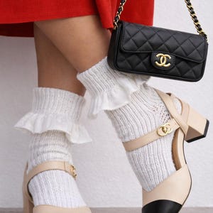 May include: A black quilted leather handbag with a gold chain strap and logo. The image also shows cream-colored Mary Jane style shoes with black toes, white socks with ruffles, and a red skirt.