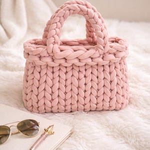 Peut inclure: Un sac fourre-tout rose clair tricoté à la main, avec une poignée tressée. Le sac est fait d'un fil épais et doux et repose sur une surface blanche et moelleuse. Des lunettes de soleil à monture dorée et un stylo sont posés sur un livre.