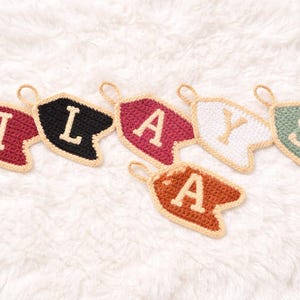 May include: Handmade crochet banner-shaped ornaments in various colors, spelling out "ILAYS". Each banner features a different color, including burgundy, black, and white, with a contrasting letter. The ornaments have a beige border and a loop for hanging.