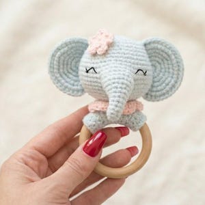 Puede incluir: Un sonajero de bebé de ganchillo con forma de elefante azul claro con una flor y un collar rosas. El elefante tiene los ojos cerrados y una larga trompa. El sonajero tiene una base de anillo de madera.