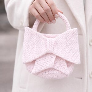 May include: A light pink crochet handbag with a bow-shaped design. The bag has a rounded base and a handle, and the bow is the focal point. The bag is held against a white coat.