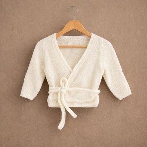 Può includere: Cardigan avvolgente color crema per neonati, con maniche lunghe e chiusura con fiocco. Il maglione è realizzato in morbido tessuto a maglia strutturata ed è appeso a una gruccia di legno. Lo sfondo è marrone.