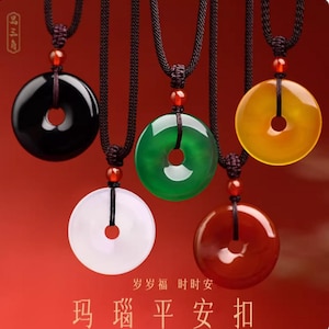 Puede incluir: Cinco collares con colgantes de jade en negro, verde, blanco, amarillo y rojo. Cada colgante es un disco circular con un agujero central, suspendido de un cordón negro. El fondo es rojo degradado.