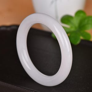 Puede incluir: Un brazalete de jade blanco liso. El brazalete circular es de un blanco opaco sólido con un acabado pulido y brillante. El fondo está borroso, con una superficie oscura y follaje verde visible.