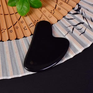 May include: A black gua sha tool with a heart-shaped design, resting on a decorative fan. The fan has a bamboo frame and a white and gray patterned surface. A green leaf is visible in the upper left corner.