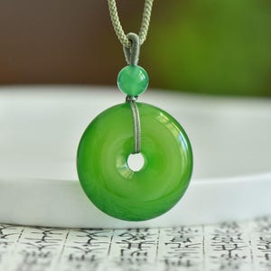 Könnte beinhalten: Eine leuchtend grüne Jade-Anhänger-Halskette. Der Anhänger ist eine kreisförmige Scheibe mit einem zentralen Loch, die an einer passenden grünen Kordel hängt. Eine kleinere Jadeperle sitzt über der Scheibe. Die Halskette wird vor einem weichen, verschwommenen Hintergrund präsentiert.