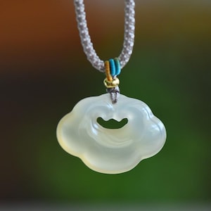 May include: A cloud-shaped pendant necklace in a pale yellow-green hue. The pendant has a curved opening and hangs from a grey, braided cord with blue and gold details. The background is a blurred mix of green and brown, creating a soft focus.
