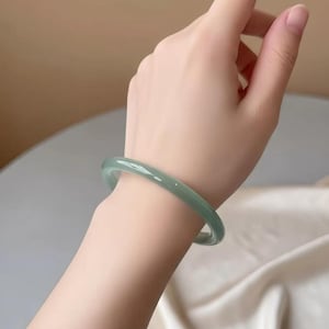 May include: A smooth, solid, light green bangle bracelet with a polished finish. The circular accessory features a translucent appearance and a rounded profile, encircling the wrist with a simple, continuous band design that lacks any ornamentation.