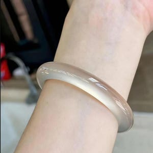 Puede incluir: Una pulsera tipo brazalete lisa, translúcida y de color beige claro con un acabado pulido. El accesorio circular presenta un perfil redondeado y un color sólido y constante, reflejando la luz en su superficie curva y brillante.