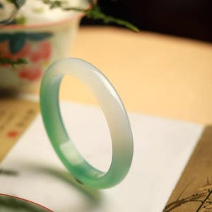 Puede incluir: Un brazalete de jade con una superficie lisa y pulida. El brazalete hace la transición de un blanco translúcido a un tono verde vibrante. El brazalete se muestra sobre una superficie blanca con un fondo borroso.