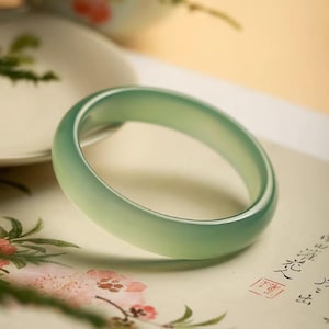 Puede incluir: Un brazalete de jade verde claro. El brazalete circular descansa sobre papel con diseños florales y caligráficos. El jade tiene una apariencia translúcida, y la estética general es elegante y refinada. El diámetro del brazalete es de aproximadamente 6,5 cm.