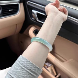 Puede incluir: Un brazalete de jade verde claro. El brazalete liso y redondeado se lleva en la muñeca. El jade tiene una cualidad translúcida, con un sutil degradado de color. El brazalete es una pieza de joyería clásica.