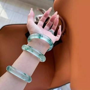 May include: Three jadeite jade bangles in varying sizes are worn on a wrist. The translucent bangles have a light green and white color with natural variations. A matching jade ring is also visible.