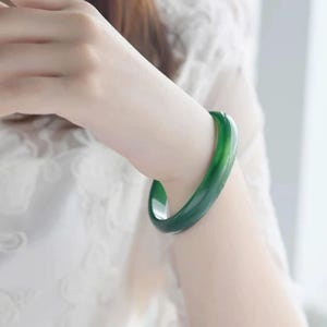 Puede incluir: Un brazalete de jade verde vibrante. La superficie lisa y pulida refleja la luz, mostrando el rico color de la piedra. Una pieza de joyería clásica y elegante.